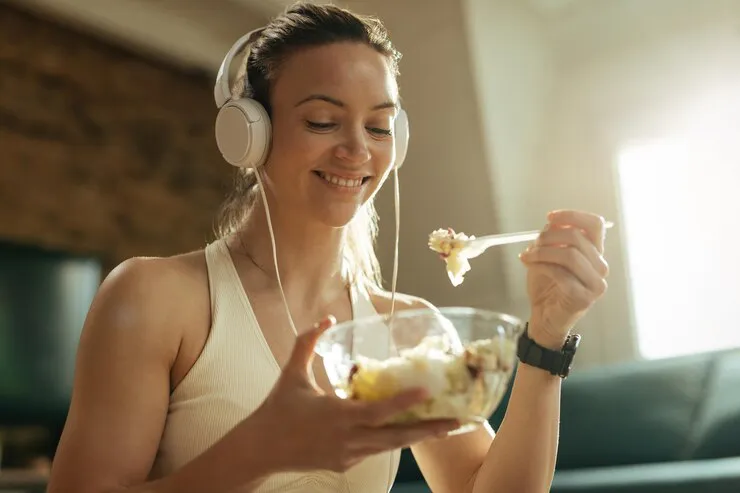 mujer-atletica-feliz-comiendo-ensalada-mientras-escucha-musica-auriculares-relaja-casa_637285-3009